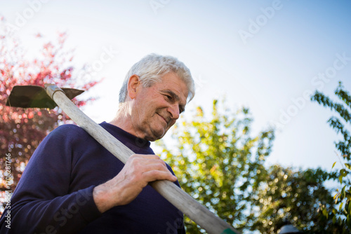 Portrait of a 70 year old man smiling in his garden with a shovel on his shoulder. Low angle view.