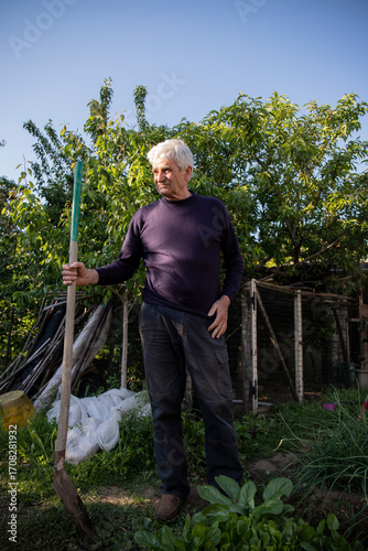 Portrait of a 70 year old man standing in his garden with a shovel.  Vertical shot. Background with copy space.
