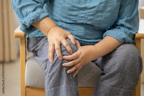 Asian Middle aged person holding knee in pain sitting on chair, discomfort and joint ache concept