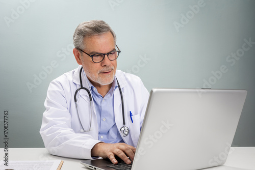 A doctor in a white coat is sitting at a desk, using a laptop to conduct an online consultation. He is engaged, offering guidance and support to a patient remotely.