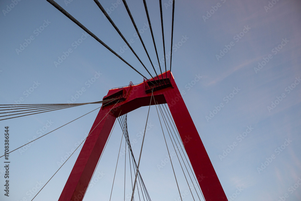 Fototapeta premium Red suspension bridge tower with cables extending towards the sky, showcasing modern engineering and architectural design against a serene evening backdrop