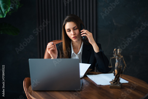 A woman is comfortably sitting at a modern desk with a laptop open in front of her while she is simultaneously talking on her cell phone