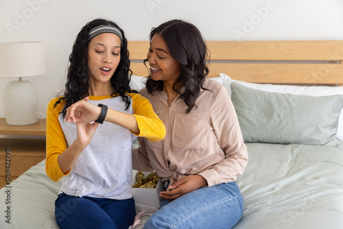 Diverse mother and daughter hugging while examining smartwatch and gift box in bedroom