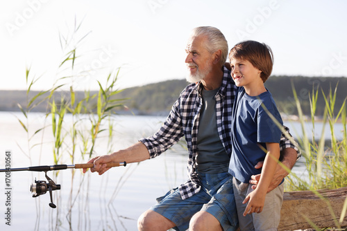 Tablou pe pânză Smiling grandfather and his nephew fishing together near river outdoors