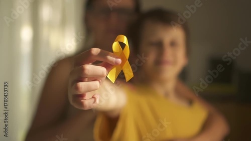 Emotional moment of mother hugging her teenage son who holds yellow satin ribbon symbolizing pediatric cancer awareness and hope.