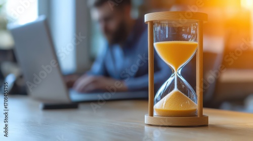 A close-up of an hourglass filled with sand, set on a desk, with a blurred figure working on a laptop in the background, symbolizing time management.