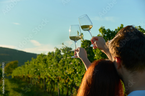 Horizontal photo of a couple with heads close together, focusing on white wine glasses. Sunlight illuminates the wine, while vineyard rows in the background are softly blurred, creating romantic and c