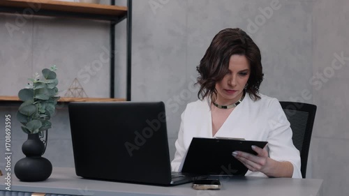 professional woman, diligent woman, corporate workspace. Professional woman in white suit works intently on paperwork at her desk, accompanied by laptop and smartphone in modern office.