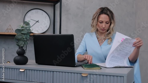 professional woman, office reading, focused woman. Young professional woman in blue outfit carefully reading and analyzing printed documents while working at modern office desk.