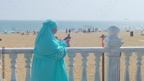 A young Muslim woman in a blue hijab stands by the sea on the embankment and works on her smartphone, business correspondence. General shot.
