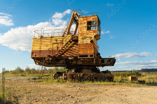 Old rusty excavator stands idle in an open field under a blue sky, showing signs of wear and abandonment in a rural industrial setting.