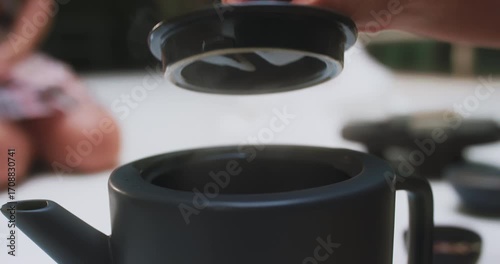 Elegant hand lifting teapot lid as steam rises, close-up
