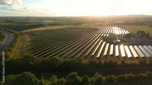 Aerial done view of solar power plant, photovoltaic panels generating renewable energy electricity in rural landscape at sunset with train passing along tracks, England UK