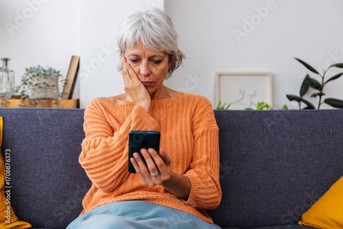 Worried senior woman reading bad news on smart phone message sitting on sofa at home.