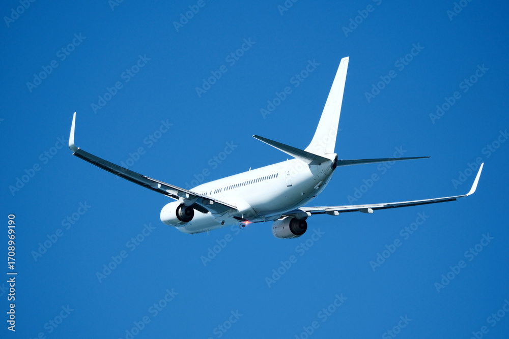 Fototapeta premium The plane that's just taken off drifts away from the airport across clear blue sky. Airplane flying in the blue sky on a sunny day. Airplane taking off, rear view. Travel concept.