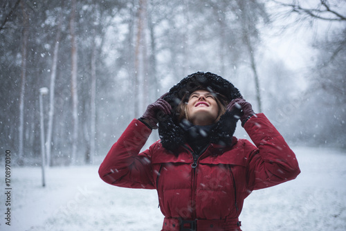 Portrait of beautiful young woman in red winter hooded jacket, while looking and and cheerful watching snowflakes falling all around. Fashion model in winter scene forest, snowing, Christmas, season.