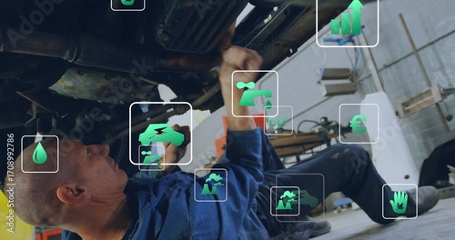 Wrenching mechanic wearing blue coverall addressing car undercarriage in workshop, with green icons