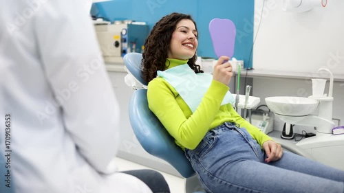 Young woman sitting in a dental chair and looking in the mirror. Happy patient smiling after a successful dental treatment or checkup