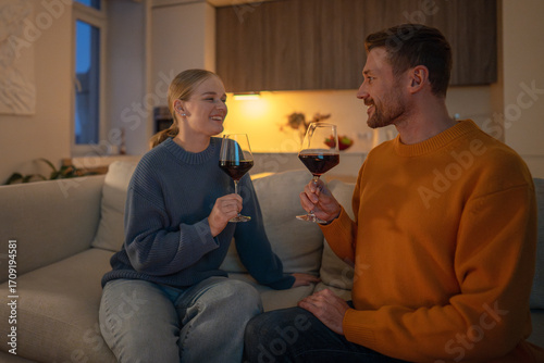 Laughing couple drinking wine on sofa looking at each other lovingly in intimate light. Romantic evening, emotional connection, love relationship, man and woman bonding at home, date on weekends