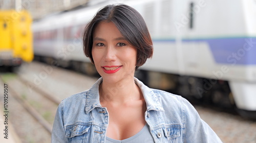 Young woman in denim jacket standing on a train platform, looking directly at the camera with blurred trains in the background