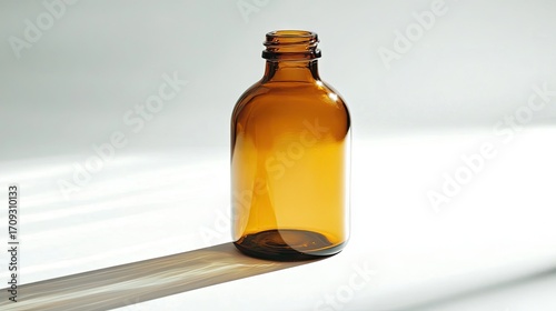 Amber glass bottle, cylindrical shape, empty, sits on a bright white surface casting a long shadow from sunlight.  The bottle's neck is slightly visible
