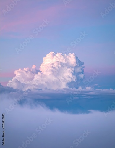 A majestic cumulus cloud formation is beautifully illuminated in soft, pastel hues during a serene sunset, floating above hazy terrain