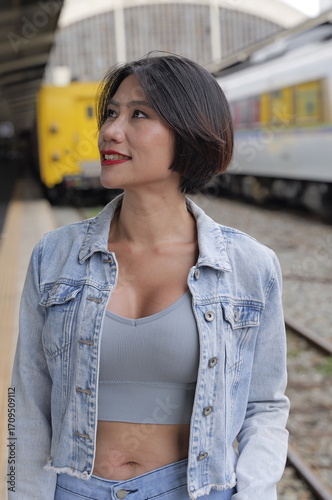 Young woman in denim jacket standing on a train platform with blurred trains in the background