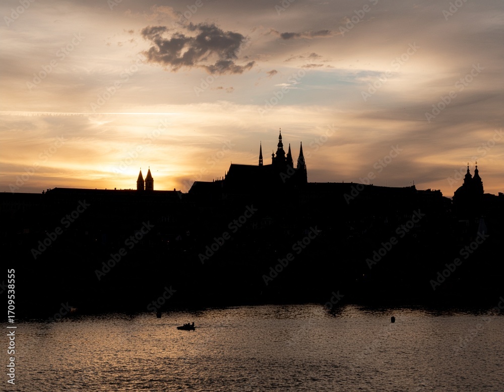 Fototapeta premium Prague Castle Silhouette at Sunset: A dramatic silhouette of Prague Castle and cityscape against a vibrant sunset sky, reflecting in the calm waters below, creating a sense of wonder and grandeur. 
