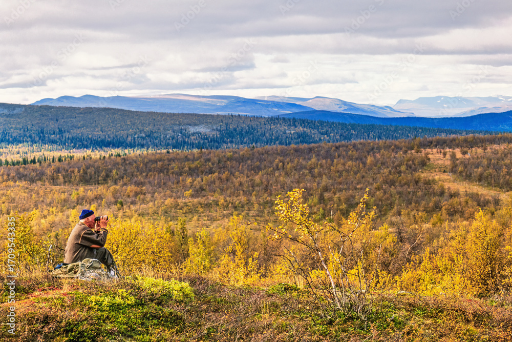 Obraz premium Man sitting and looking through binoculars in a mountain landscape in autumn