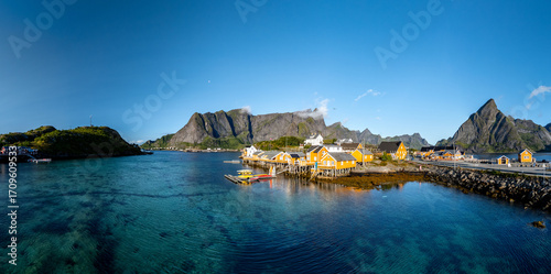 Brightly painted cabins line the tranquil waters of Sakrisoy, Lofoten, Norway, under a clear blue sky.