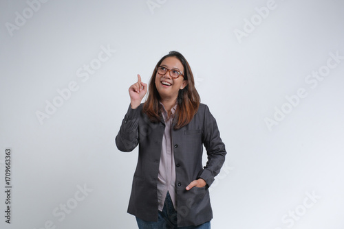 An Asian businesswoman wearing glasses and a blazer with a cheerful expression,one hand pointing up. Studio shot with white background.