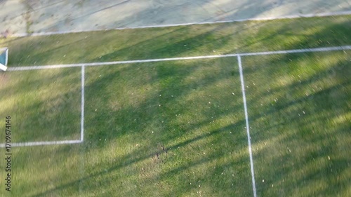 Aerial view of a football pitch corner and goal box with long evening shadows on green grass. Minimal, geometric sports field lines. No people.
