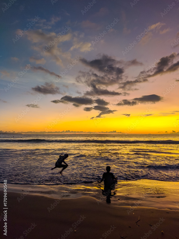 Obraz premium Golden sunset paints the sky while two kids create playful silhouettes along the gentle waves of Kuta Beach, capturing carefree joy as colorful reflections shimmer across the wet shoreline.
