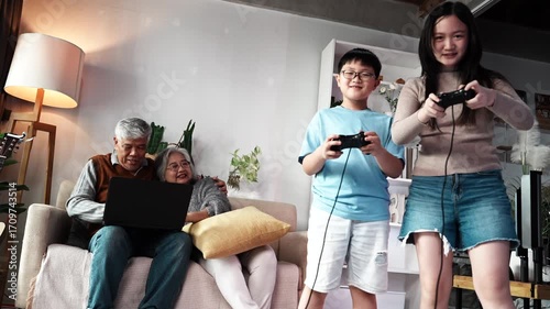 An Asian family enjoys playing eSports at home with grandparents cheering on their grandchildren on the sofa. The granddaughter and her teenage brother and sister hold joysticks and play games.