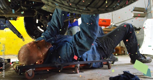 Mechanic lying on creeper inspecting suspension under raised vehicle at garage, drifting particles