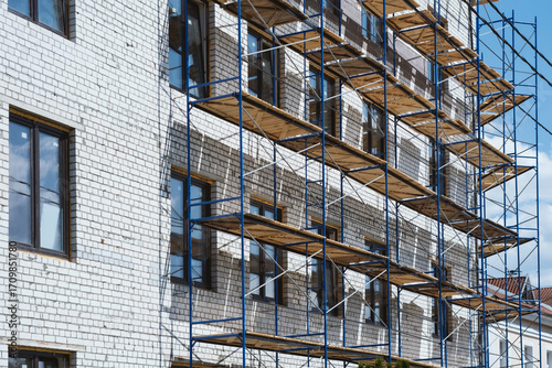 Multi-level blue metal scaffolding with wooden platforms erected against white brick apartment building under construction.