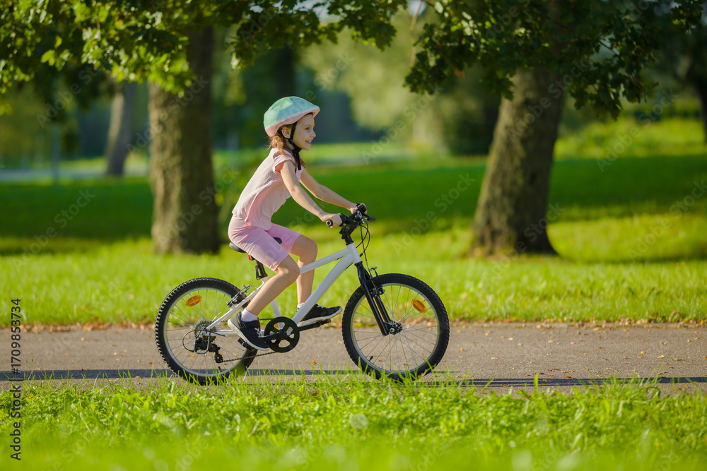 Obraz premium Happy smiling little girl with helmet riding on white bike on road at town park in warm summer day. Enjoying cycling. Cute 6 years old child. Side view.