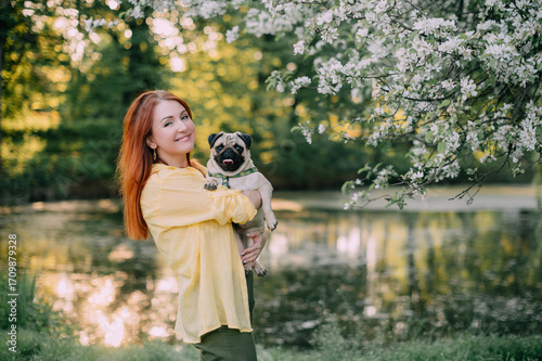 A young redhead woman holds her pug dog in front of her. The owner and the pet are having fun outdoors. 