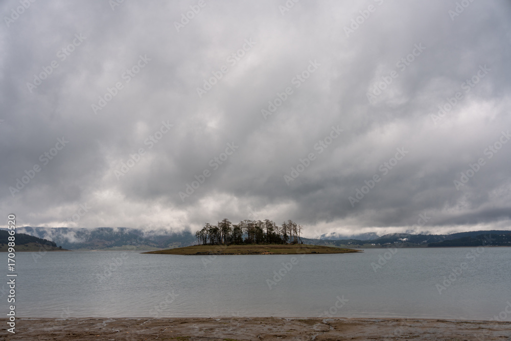 Fototapeta premium Moody autumn view of Batak Reservoir in Rhodope Mountains, Bulgaria, with small island under gray cloudy sky, scenic spot for eco tourism and nature travel