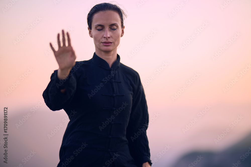 Fototapeta premium Woman practicing Tai Chi in traditional black outfit on mountain at sunrise, arms outstretched in meditation pose focusing on balance, energy, and mindfulness in nature.