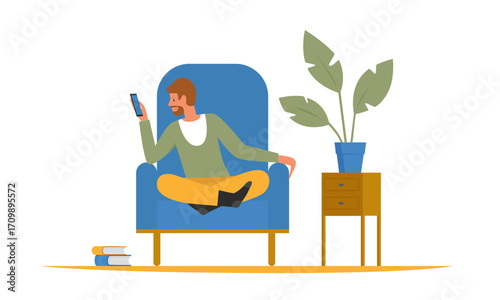 A man sitting in a blue armchair, looking at his phone, with a plant and side table.