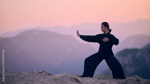 Foto Woman practicing Tai Chi in traditional black outfit on mountain at sunrise, arms outstretched in meditation pose focusing on balance, energy, and mindfulness in nature