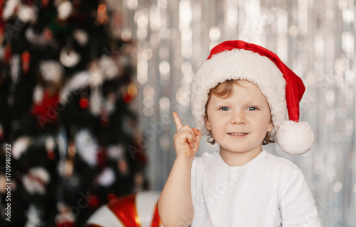 A little boy in a Santa hat smiles happily in front of festive Christmas decorations, with twinkling lights and ornaments creating a joyful holiday atmosphere.