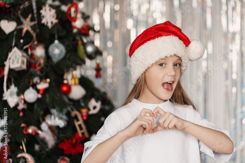 Cheerful 9-year-old girl in Santa hat making heart shape with fingers in front of decorated Christmas tree, festive holiday mood and joy