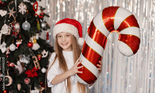 Cheerful 9-year-old girl in Santa Claus hat holding foil balloon shaped like candy cane in front of decorated Christmas tree, festive holiday mood