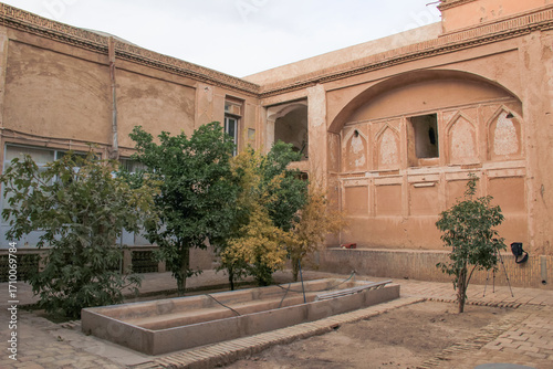 Traditional courtyard with trees and old adobe walls in Yazd, Iran, showcasing Persian architecture and desert lifestyle.