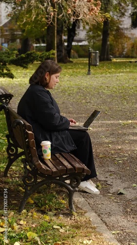A young woman sits on a park bench working on her laptop with a coffee cup beside her, enjoying a sunny autumn day. Side view portrait. Digital nomad concept. Vertical 4k footage