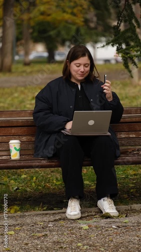 Woman sitting on a wooden park bench, smiling while talking on the phone, with her laptop and a takeaway coffee cup beside her. Digital nomad concept. Vertical 4k footage