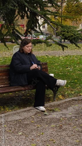 Woman sitting on a park bench writing notes in a notebook while checking her phone. Autumn leaves around create a calm outdoor work setting. Vertical 4k footage