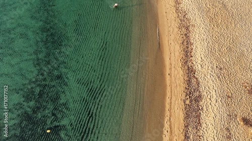 Stunning aerial drone view of a serene beachscape featuring crystal-clear greenish-blue waters gently meeting golden sand. The shoreline forms elegant wave patterns, while neatly arranged white beach 